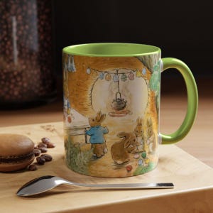 Puede incluir: Taza de cerámica con asa e interior verdes, con una ilustración caprichosa de conejos en un entorno forestal. El diseño incluye un conejo con chaqueta azul, una olla y huevos de Pascua. La taza está sobre una superficie de madera.