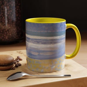 May include: A ceramic coffee mug with a yellow handle and interior. The exterior features a watercolor design in shades of blue, white, and yellow. A silver spoon, a macaron, and coffee beans are on a wooden surface.