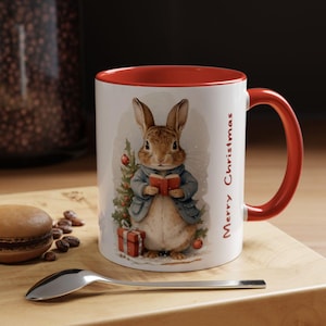 Könnte beinhalten: Weißer Keramikbecher mit rotem Innenraum und Henkel. Der Becher zeigt eine Illustration eines Hasen in einem blauen Mantel, der ein Buch hält, mit einem Weihnachtsbaum und einem Geschenk. Die Worte "Merry Christmas" sind vertikal gedruckt.