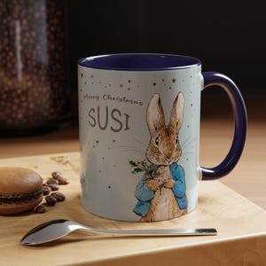 Könnte beinhalten: Ein weißer Keramikbecher mit dunkelblauem Innenraum und Henkel, verziert mit einer Aquarellillustration eines braunen Hasen in blauer Jacke, der Stechpalmen hält. Der Text Merry Christmas und der Name SUSI sind in Braun aufgedruckt.