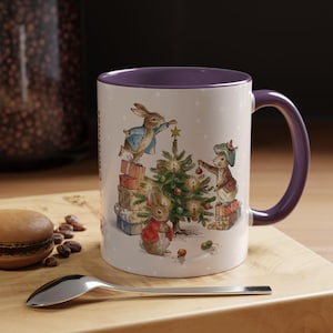 Op de afbeelding: Een witte keramische mok met een paarse binnenkant en handvat, voorzien van een aquarelillustratie van drie konijnen die een kerstboom versieren met cadeautjes, kerstballen en een ster, op een witte achtergrond met subtiele witte stippen.