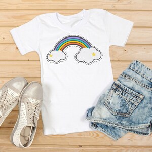 Puede incluir: Camiseta blanca con un dise&ntilde;o de arco&iacute;ris y nubes, combinada con shorts de mezclilla y zapatillas plateadas. El dise&ntilde;o del arco&iacute;ris presenta un arco colorido sobre dos nubes blancas con estrellas amarillas. El conjunto se muestra sobre una superficie de madera.