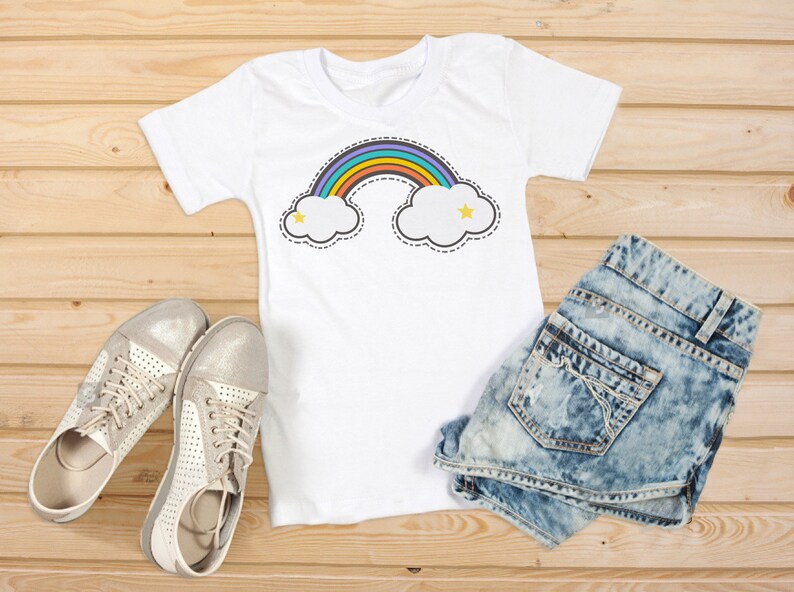 Puede incluir: Camiseta blanca con un dise&ntilde;o de arco&iacute;ris y nubes, combinada con shorts de mezclilla y zapatillas plateadas. El dise&ntilde;o del arco&iacute;ris presenta un arco colorido sobre dos nubes blancas con estrellas amarillas. El conjunto se muestra sobre una superficie de madera.
