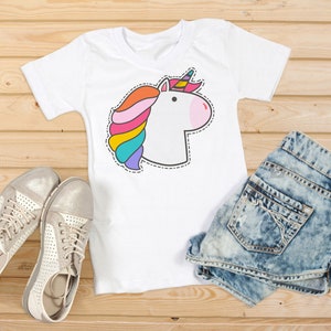 Puede incluir: Camiseta blanca con un dise&ntilde;o de unicornio colorido, con melena arco&iacute;ris y nariz rosa. La imagen incluye pantalones cortos de mezclilla y zapatillas plateadas, todo sobre una superficie de madera.