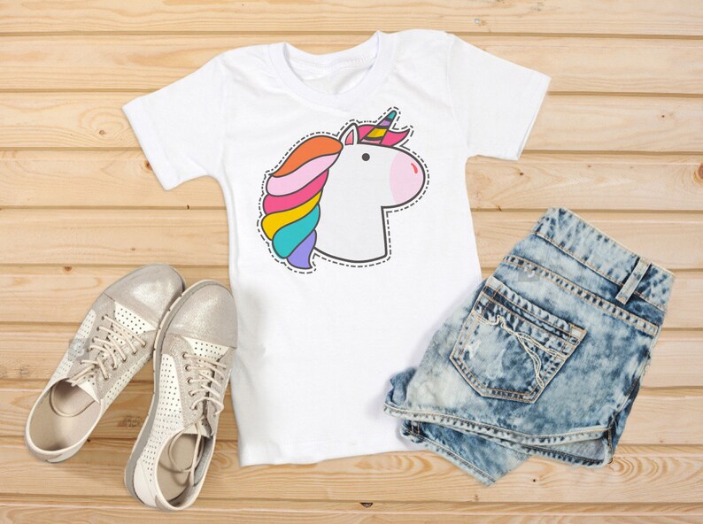 Puede incluir: Camiseta blanca con un dise&ntilde;o de unicornio colorido, con melena arco&iacute;ris y nariz rosa. La imagen incluye pantalones cortos de mezclilla y zapatillas plateadas, todo sobre una superficie de madera.