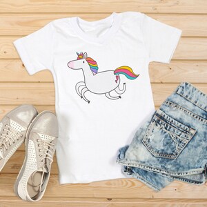 Puede incluir: Camiseta blanca con un dise&ntilde;o de unicornio de dibujos animados. El unicornio tiene una melena y cola de arco&iacute;ris. La camiseta se combina con pantalones cortos de mezclilla y zapatillas plateadas. Los art&iacute;culos est&aacute;n colocados sobre una superficie de madera.