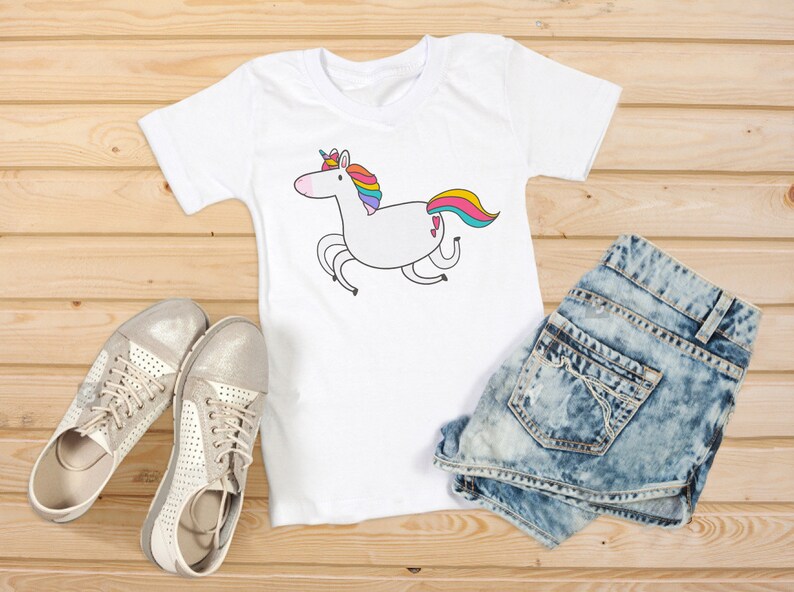 Puede incluir: Camiseta blanca con un dise&ntilde;o de unicornio de dibujos animados. El unicornio tiene una melena y cola de arco&iacute;ris. La camiseta se combina con pantalones cortos de mezclilla y zapatillas plateadas. Los art&iacute;culos est&aacute;n colocados sobre una superficie de madera.