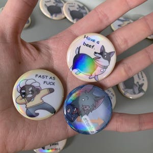 May include: A collection of round, holographic-effect buttons. One button features a cartoon corgi with the text "Have a beef." Another shows a cartoon figure dabbing with the text "Fast as Fuck." The third button has a cartoon bat with the word "Stonks."