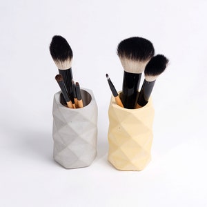 May include: Two geometric concrete makeup brush holders, one gray and one yellow, with makeup brushes inside.