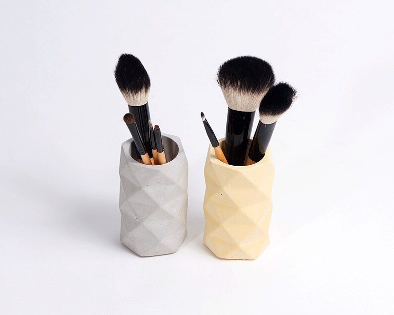 May include: Two geometric concrete makeup brush holders, one gray and one yellow, with makeup brushes inside.