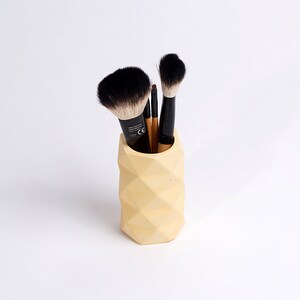 May include: A yellow geometric concrete makeup brush holder with black and brown makeup brushes inside.