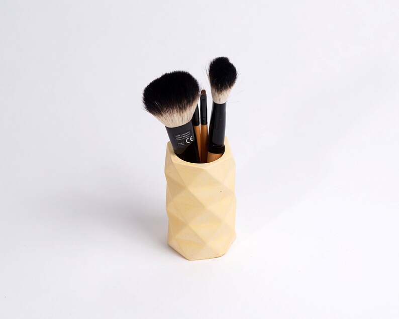 May include: A yellow geometric concrete makeup brush holder with black and brown makeup brushes inside.