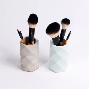 May include: Two geometric concrete makeup brush holders, one gray and one light blue, each filled with makeup brushes.
