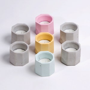May include: Eight small, octagonal concrete candle holders in various colours, including grey, light blue, yellow, and pink. Each holder has a small, white candle inside.