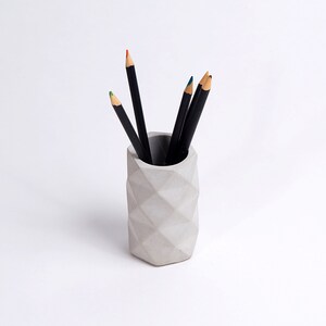 May include: A gray concrete pencil holder with a geometric design. The holder is filled with black pencils.