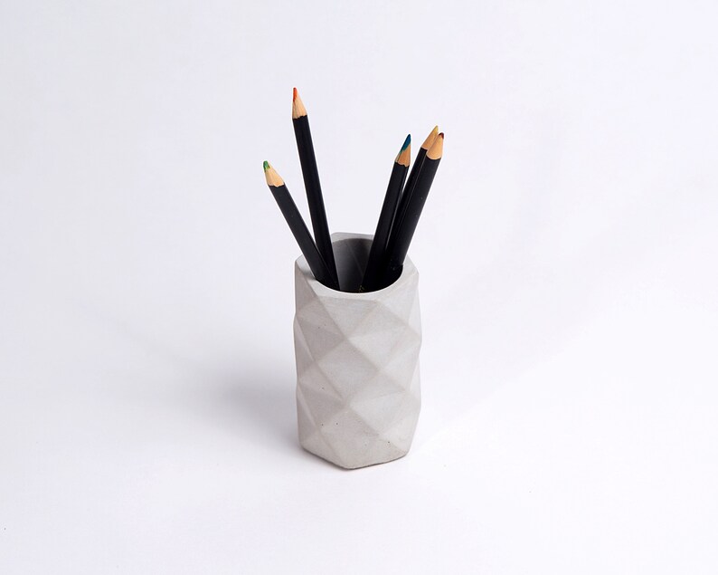 May include: A gray concrete pencil holder with a geometric design. The holder is filled with black pencils.