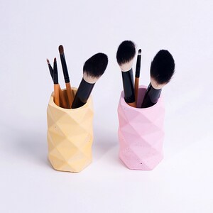 May include: Two geometric concrete makeup brush holders, one yellow and one pink, with makeup brushes inside.