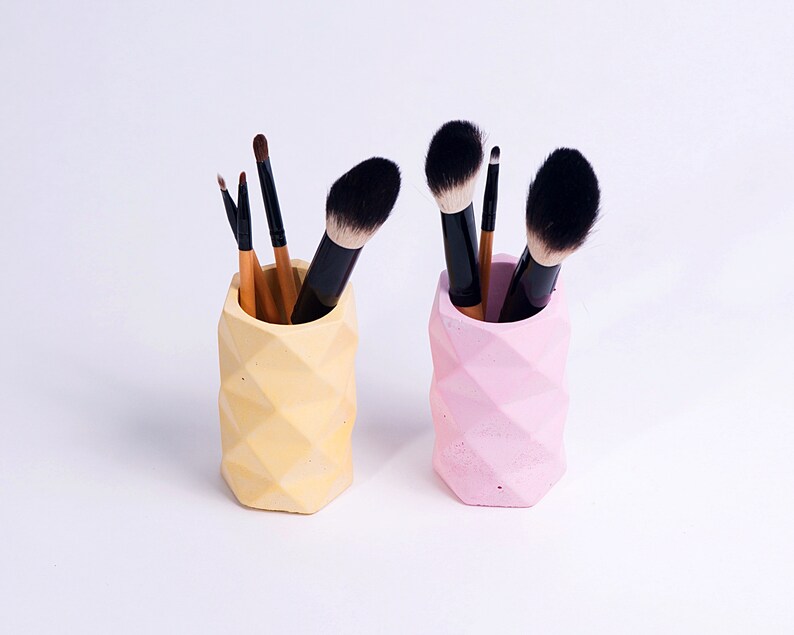May include: Two geometric concrete makeup brush holders, one yellow and one pink, with makeup brushes inside.