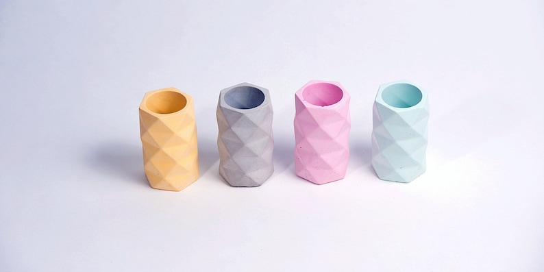 May include: Four geometric concrete planters in different colors: yellow, gray, pink, and light blue. The planters are all the same size and shape, with a hexagonal base and a cylindrical body.