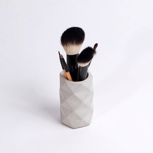 May include: A gray concrete makeup brush holder with a geometric design. Five makeup brushes are in the holder, including a large, fluffy brush.