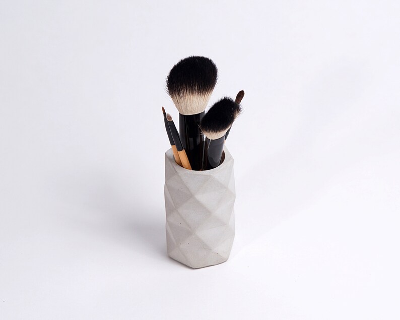 May include: A gray concrete makeup brush holder with a geometric design. Five makeup brushes are in the holder, including a large, fluffy brush.