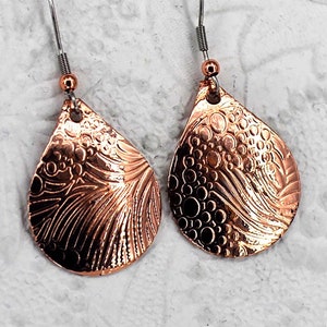 May include: A pair of copper teardrop earrings with a textured design. The earrings have a hammered finish and a delicate, intricate pattern.