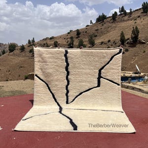 Puede incluir: Una alfombra color crema con un diseño geométrico de líneas negras. La alfombra se exhibe al aire libre contra un telón de fondo de un paisaje montañoso bajo un cielo nublado. El texto "TheBerberWeaver" es visible.