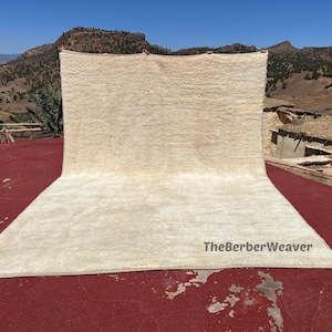 Puede incluir: Una alfombra grande, rectangular, de color crema con una superficie texturizada se exhibe al aire libre. La alfombra está colocada sobre una superficie roja, con un paisaje montañoso al fondo. El texto "TheBerberWeaver" es visible en la alfombra.