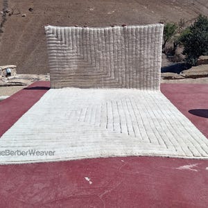 Puede incluir: Una alfombra blanca tejida con un patrón geométrico. La alfombra está colocada sobre una superficie roja. El texto "TheBerberWeaver" es visible en la esquina inferior izquierda de la alfombra.