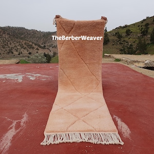 Puede incluir: Una alfombra rectangular de color melocotón con un patrón de diamantes y flecos blancos. La alfombra se exhibe al aire libre sobre una superficie roja, con el texto "TheBerberWeaver" visible en la alfombra.