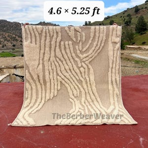 Può includere: Un tappeto beige con un motivo astratto color crema che ricorda la corteccia degli alberi. Il tappeto misura circa 140 x 160 cm ed è tenuto contro uno sfondo montuoso. Il testo "TheBerberWeaver" è visibile.