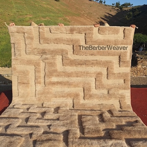 May include: A beige rug with a geometric pattern is held up against a natural backdrop. The rug features a textured design with raised rectangular and linear elements in varying shades of beige and brown. The text "TheBerberWeaver" is visible on the rug.