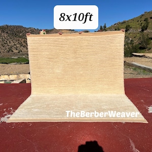 Può includere: Un tappeto rettangolare beige, di circa 244 x 305 cm, è esposto all'aperto. Il tappeto ha un motivo strutturato e a righe. Il testo "8x10ft" è in una casella bianca in alto e "TheBerberWeaver" in basso.