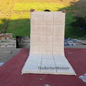 Pode incluir: Um tapete retangular de cor branco-sujo com uma grelha de dezasseis secções quadradas tufadas. O tapete apresenta o texto The Berber Weaver num logótipo quadrado preto à esquerda e o texto TheBerberWeaver impresso na secção inferior direita.