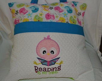 Pode incluir: Uma almofada branca com uma alça azul e uma estampa de pássaros coloridos nas costas. A frente da almofada tem um pássaro rosa bordado segurando um livro com o texto "Reading is my thing".