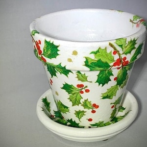 May include: A white ceramic flower pot with a matching saucer. The pot is decorated with a pattern of green holly leaves and red berries. Gold dots are scattered throughout the design. The rim of the pot is also decorated with the holly and berry pattern.