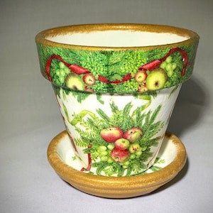 May include: A white ceramic planter with a gold rim and a festive Christmas design featuring red berries, green leaves, and apples. The planter has a matching saucer.