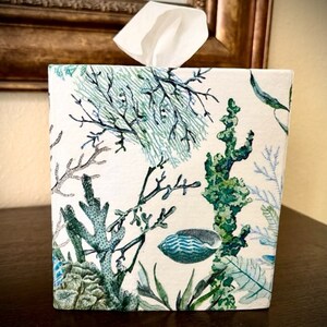 Pu&ograve; includere: Un copri scatola di fazzoletti bianco con un motivo di vita marina verde e blu. Il motivo presenta coralli, alghe e conchiglie.