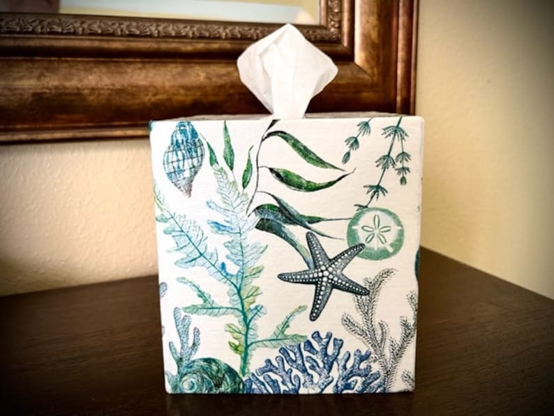 Pu&ograve; includere: Un copri scatola di fazzoletti bianco con un design costiero che presenta alghe verdi, stelle marine blu e conchiglie.