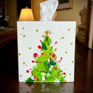 May include: White square tissue box with a hand-painted Christmas tree design. The tree is green with red and gold accents, and gold dots are scattered across the white background. A bundle of white tissues is visible at the top.