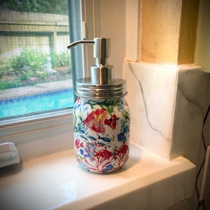 May include: A white ceramic soap dispenser with a silver pump and a floral design featuring red, pink, blue, and green flowers and fish. The dispenser is sitting on a white countertop.
