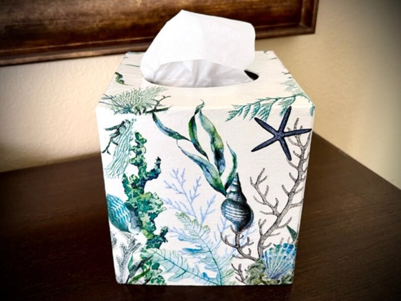 Pu&ograve; includere: Un copri scatola di fazzoletti bianco con un design a tema marino. Il design presenta stelle marine blu, alghe verdi e conchiglie bianche su uno sfondo bianco.