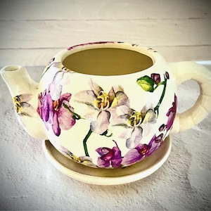 May include: Cream ceramic teapot with a matching saucer, decorated with a floral orchid design in purple, white, and green. The teapot has a rounded body, a curved handle, and a spout.