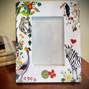 May include: White picture frame decorated with illustrations of a monkey, toucan, zebra, and crane, alongside flowers and leaves. The frame has a rectangular aperture for a photograph.