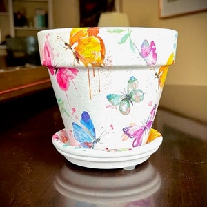 Könnte beinhalten: Ein weißer Keramik-Blumentopf mit passender Untertasse, verziert mit bunten Aquarell-Schmetterlingen in Rosa, Gelb, Blau und Grün. Der Topf hat eine leicht ausgestellte Form und ist für den Innen- oder Außenbereich geeignet.