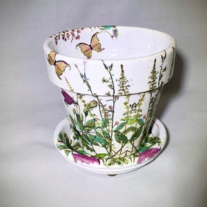 Puede incluir: Maceta de cerámica blanca con un platillo. La maceta y el platillo están decorados con un patrón floral con mariposas, hojas verdes y flores rosas.