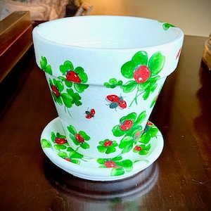 Puede incluir: Maceta de cerámica blanca con un diseño de trébol verde y mariquita. La maceta tiene un platillo a juego.