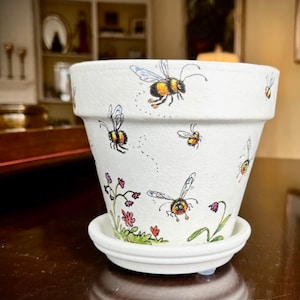 May include: White ceramic flower pot with a painted design of bees and flowers. The pot has a saucer.