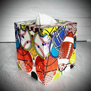 Könnte beinhalten: Ein quadratischer Taschentuchspender mit einer Collage zum Thema Sport. Das Design umfasst Basebälle, Basketbälle und einen Football, mit einer Mischung aus Farben und Mustern. Weiße Taschentücher sind oben sichtbar.