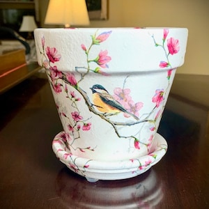 May include: White ceramic flower pot with a pink and white floral design and a small bird perched on a branch. The pot has a matching saucer.